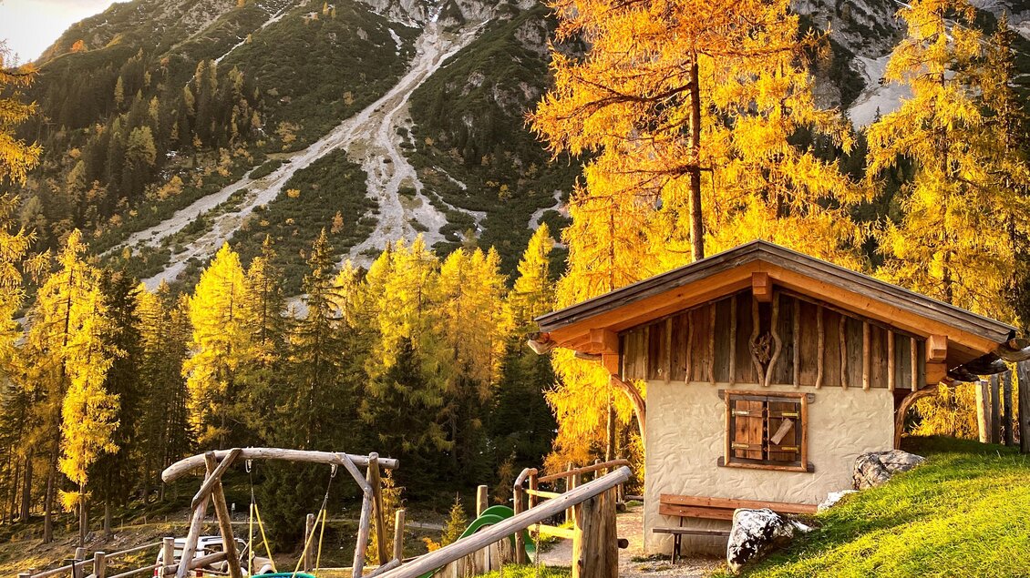 Eine malerische Almhütte umgeben von bunten Lärchen im Herbst. Im Hintergrund erheben sich beeindruckende Berge unter einem bewölkten Himmel. | © ©KajetanGerharter 