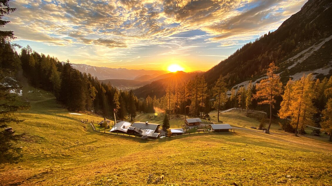 Ein malerischer Sonnenuntergang über einer Berglandschaft mit sanften Hügeln und vereinzelten Bäumen. Im Vordergrund sind einige kleine Hütten zu sehen. | © ©KajetanGerharter 