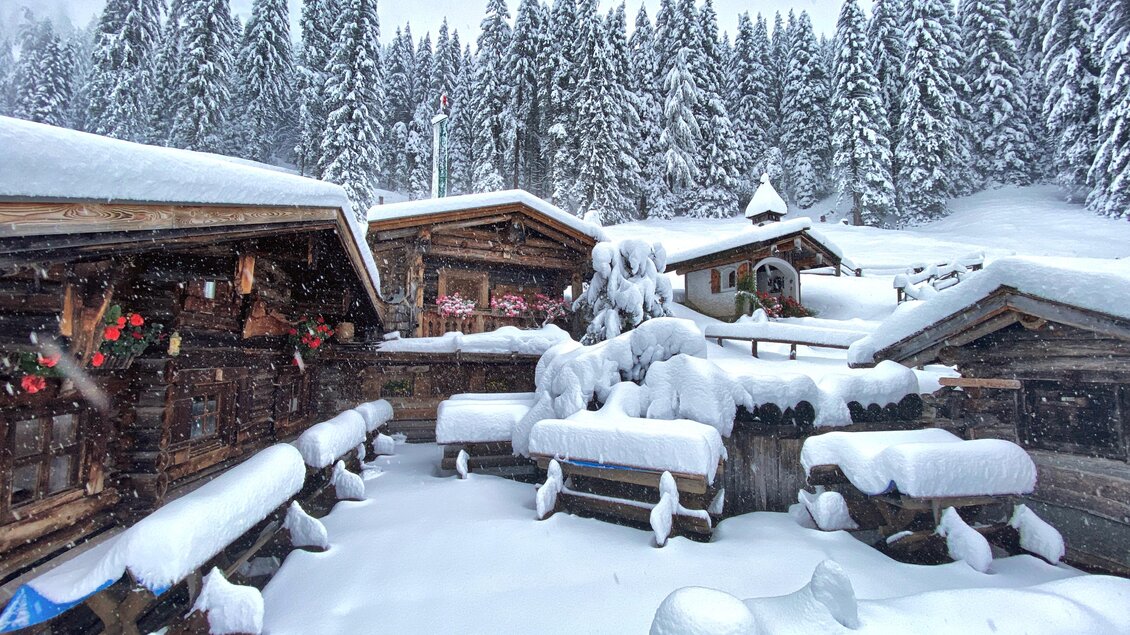Eine verschneite Winterlandschaft mit Holzblockhäusern und einem dichten Wald im Hintergrund. Der Schnee bedeckt alles und schafft eine idyllische, ruhige Atmosphäre. | © ©KajetanGerharter 