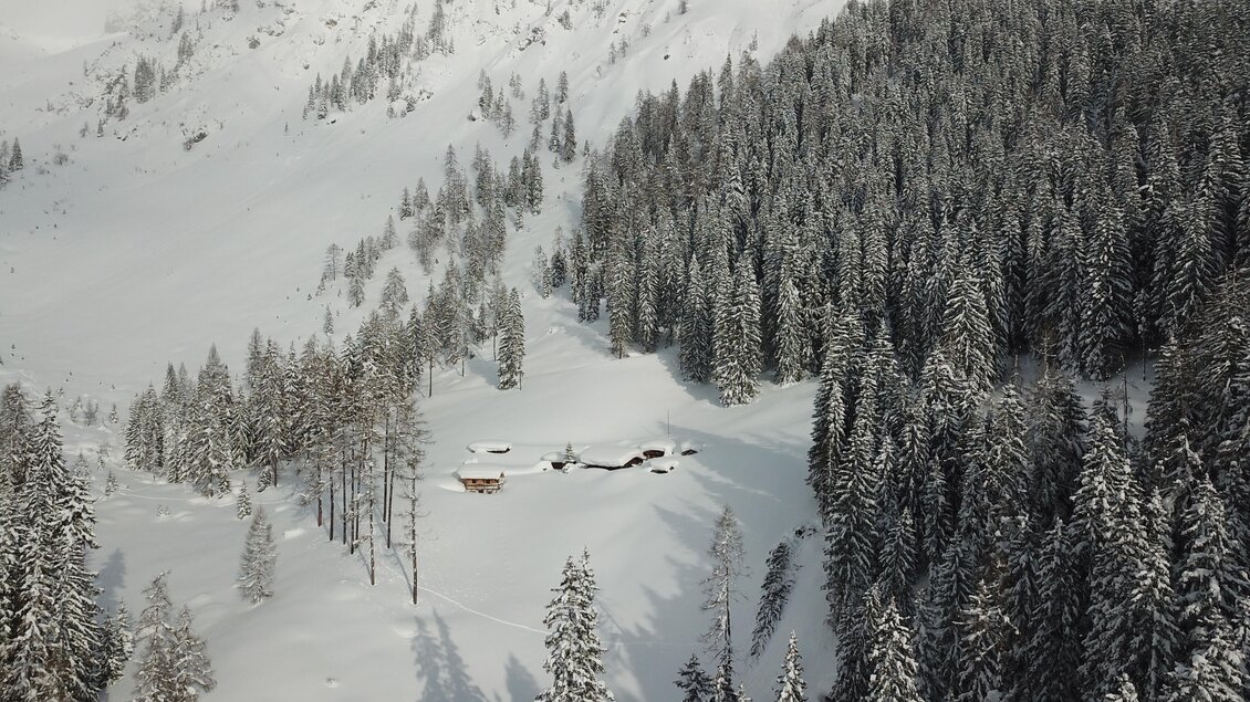Eine verschneite Landschaft mit hohen Tannen und einem Berg im Hintergrund. In der Mitte sind einige Hütten sichtbar, umgeben von weißem Schnee. | © ©KajetanGerharter 
