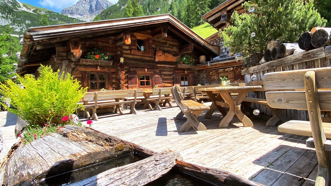 Eine gemütliche Alphütte mit Holzterrasse und Blick auf die Berge. Umgeben von Natur und Pflanzen, fließt klares Wasser in einem kleinen Teich. | © ©KajetanGerharter 
