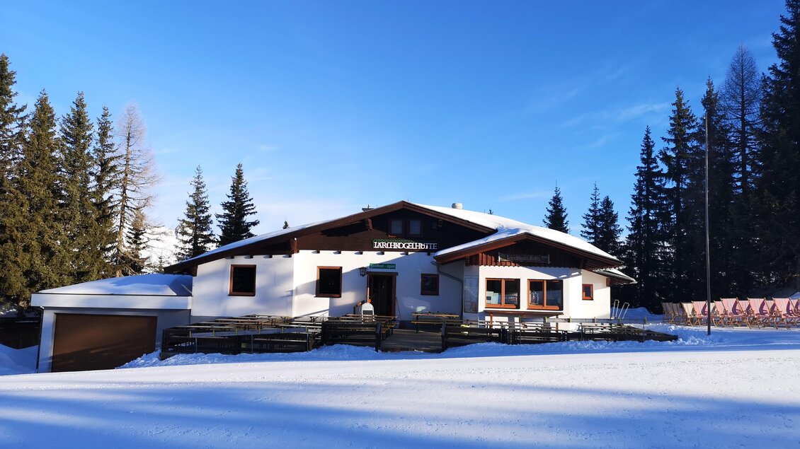 Ein gemütliches, weißes Haus im Schnee, umgeben von hohen Tannen. Der klare blaue Himmel sorgt für eine strahlende Winterstimmung. | © Lärchkogelhütte