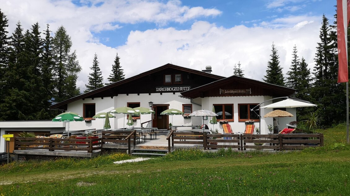 Eine gemütliche Berghütte umgeben von Bäumen und einer grünen Wiese. Auf der Terrasse stehen Sonnenschirme und Sitzgelegenheiten. | © Lärchkogelhütte