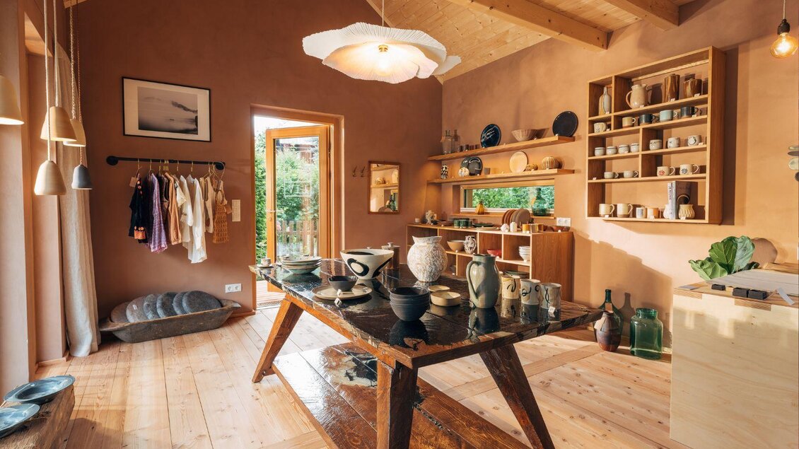 Ein stilvolles Atelier mit warmen Erdtönen und Holzfußboden. Auf einem großen Tisch stehen Keramiken und Handwerkstücke, während einige Kleidungsstücke an der Wand hängen. | © Zinnebener Lisa