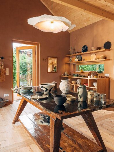 Ein stilvolles Atelier mit warmen Erdtönen und Holzfußboden. Auf einem großen Tisch stehen Keramiken und Handwerkstücke, während einige Kleidungsstücke an der Wand hängen. | © Zinnebener Lisa
