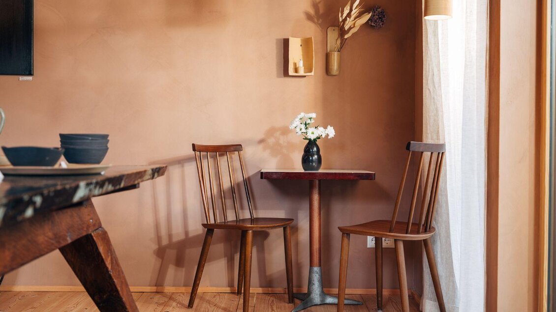 Ein gemütlicher Raum mit zwei Holzstühlen und einem kleinen Tisch. Eine Vase mit weißen Blumen steht auf dem Tisch vor einer warmen, erdigen Wandfarbe. | © Lisa Zinnebner