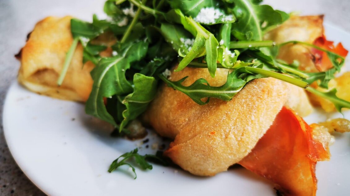 Ein Teller mit frisch gebackenem Teig, gefüllt mit Schinken und Rucola. Das Gericht sieht appetitlich und köstlich aus. | © La Piazzetta