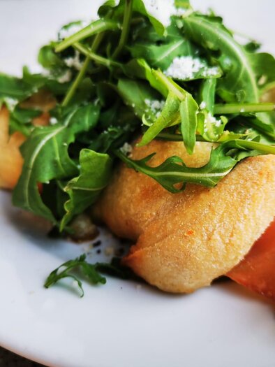 A plate with freshly baked dough, filled with ham and arugula. The dish looks appetizing and delicious. | © La Piazzetta