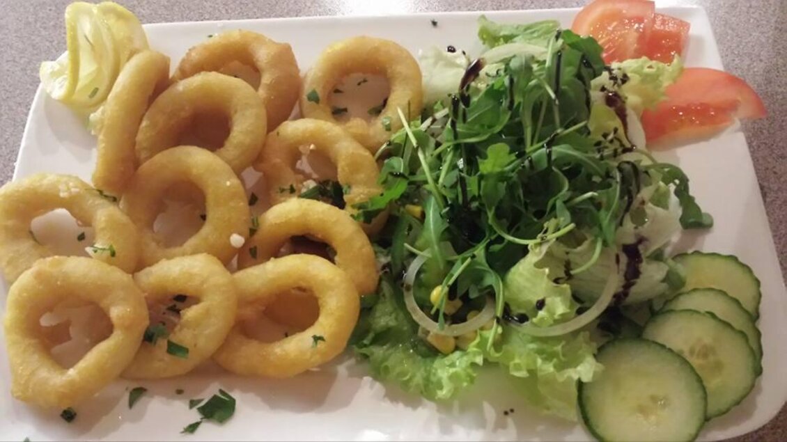 Ein Teller mit knusprigen Calamari-Ringen und buntem Salat. Dazu sind Gurkenscheiben, Tomaten und ein Zitronenschnitz angerichtet. | © La Piazzetta