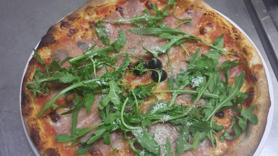 Eine frisch gebackene Pizza mit Schinken, Oliven und frischem Rucola. Der Käse ist geschmolzen und die Kruste ist goldbraun. | © La Piazzetta
