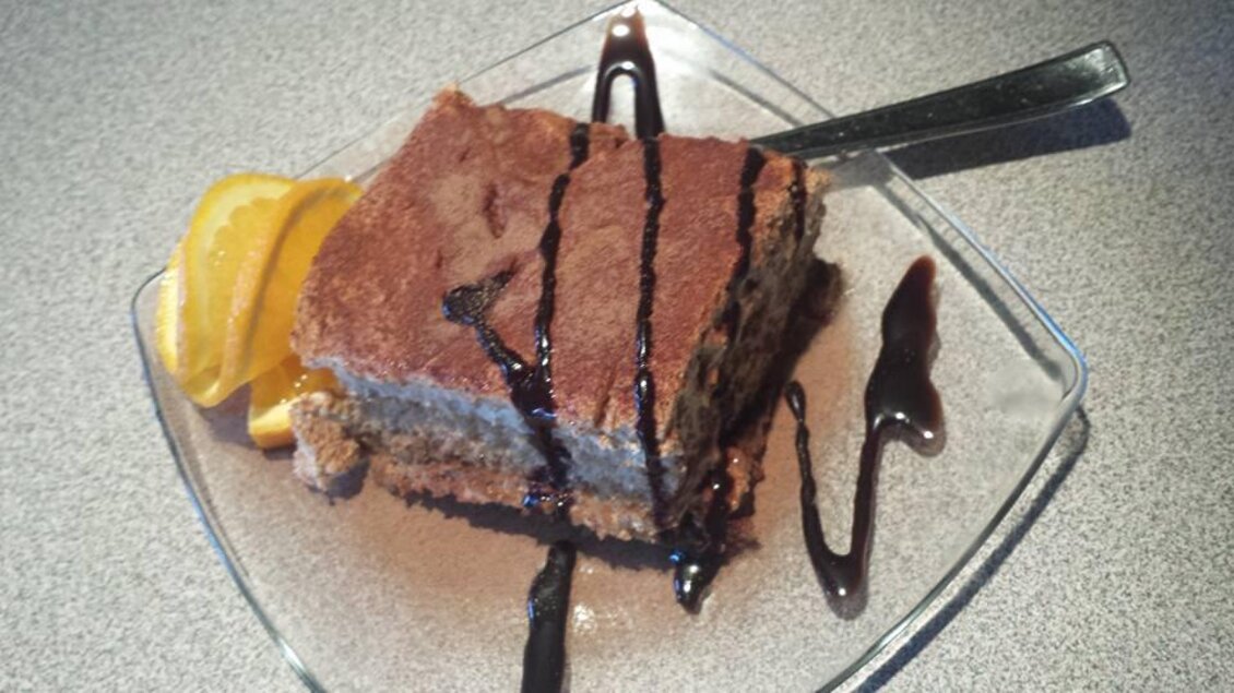 Ein Stück Tiramisu auf einem Glas-Teller, garniert mit Schokoladensauce und einer Orangenscheibe. Das Dessert sieht cremig und verführerisch aus. | © La Piazzetta