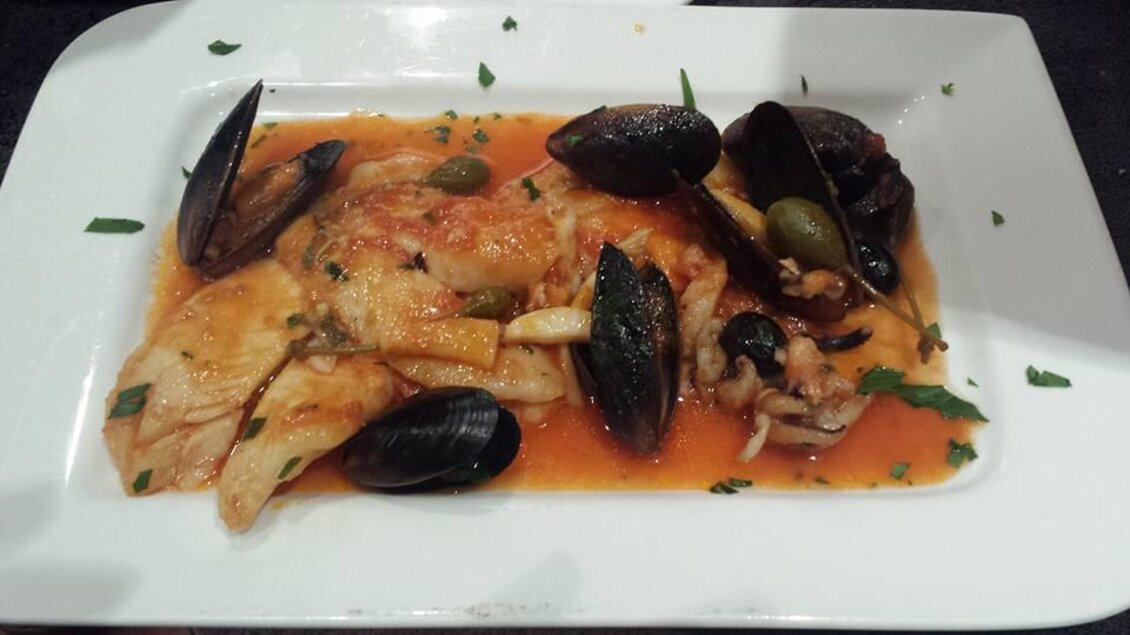 Ein Teller mit Fischfilet, Muscheln und einer würzigen Tomatensauce. Das Gericht ist mit frischen Kräutern garniert. | © La Piazzetta