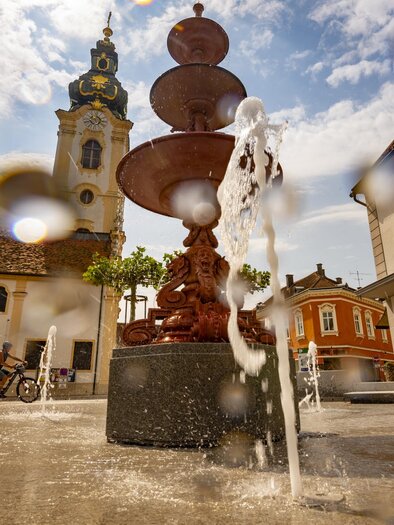 A beautiful fountain in a city with bubbling water. In the background, historical buildings and a church steeple are visible. | © Oststeiermark Tourismus