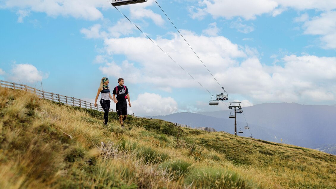 Ein Paar geht auf einem grünen Hang mit einer Seilbahn im Hintergrund. Der Himmel ist teilweise bewölkt und die Landschaft wirkt einladend. | © Lachtal-Lifte u. Seilbahnen GmbH