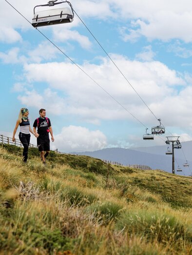 A couple is walking on a green hillside with a cable car in the background. The sky is partly cloudy and the landscape looks inviting. | © Lachtal-Lifte u. Seilbahnen GmbH
