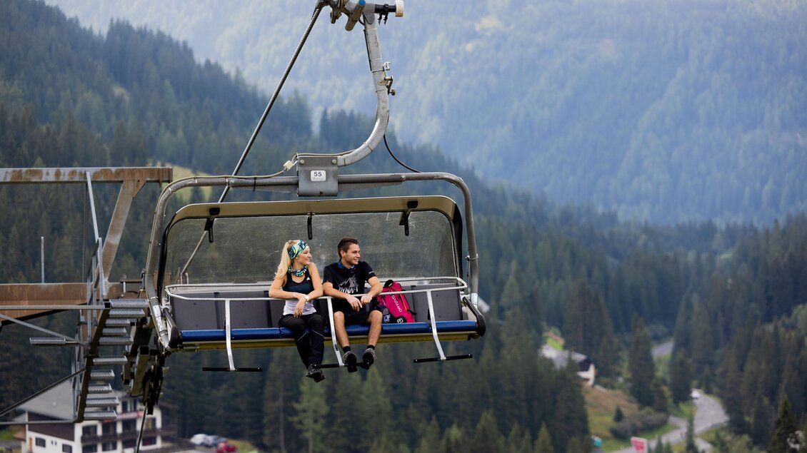 Eine Seilbahn mit zwei Personen schwebt über eine grüne Waldlandschaft. Im Hintergrund sind Berge und ein kleiner Weg sichtbar. | © Tourismusverband Murau