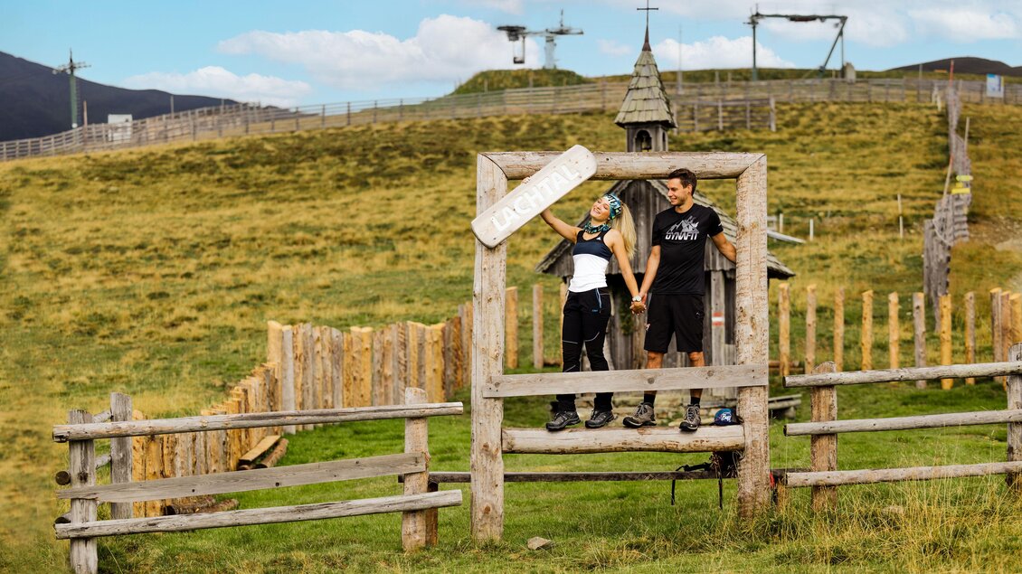 Ein Paar steht auf einer Holzstruktur in einer grünen, offenen Landschaft. Im Hintergrund sind Hütten und eine Bergkulisse zu sehen. | © Tourismusverband Murau