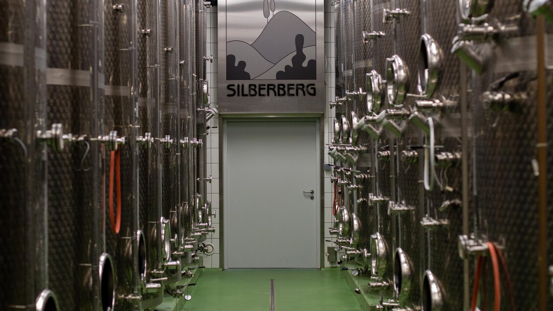Ein schmaler Gang zwischen großen, silbernen Weinfässern. Am Ende des Ganges befindet sich eine Tür mit dem Logo "Silberberg". | © @Silberberg