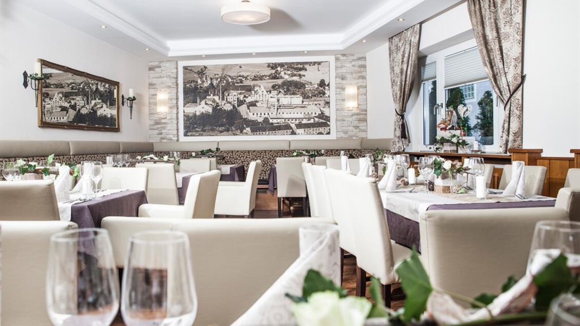 Ein eleganter Restaurantbereich mit weißen Tischen und Stühlen. Die Wände sind mit Kunstwerken dekoriert, und die Atmosphäre ist einladend und gemütlich. | © Landgasthof Erlsbacher