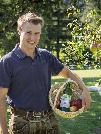 A young man is standing in a meadow holding a basket full of apples and drinks. In the background, apple trees can be seen. | © Netzwerk Kulinarik wildbild