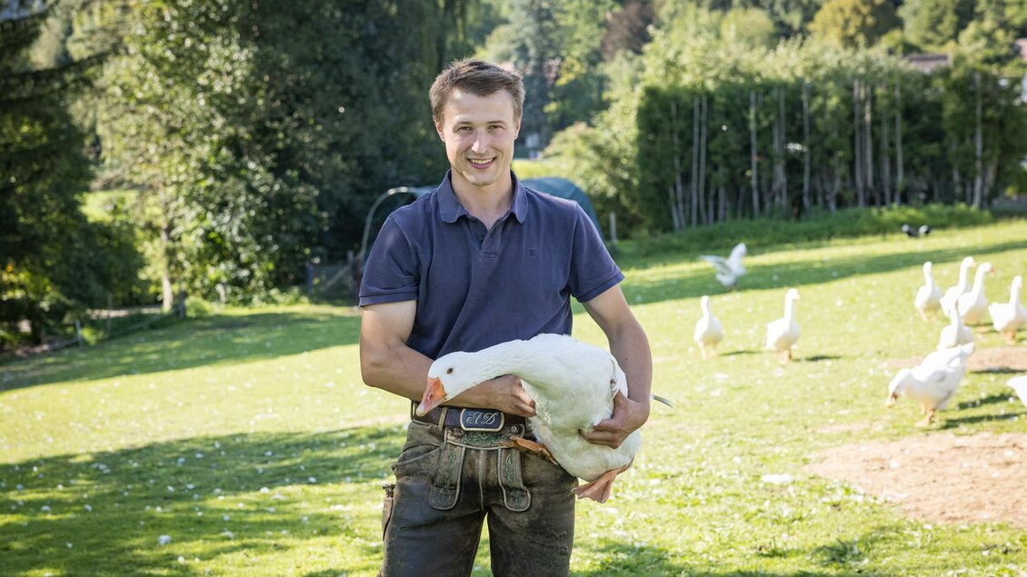 Ein junger Mann steht auf einer grünen Wiese und hält eine weiße Gans in seinen Armen. Im Hintergrund sind weitere Gänse und Bäume zu sehen. | © Netzwerk Kulinarik wildbild