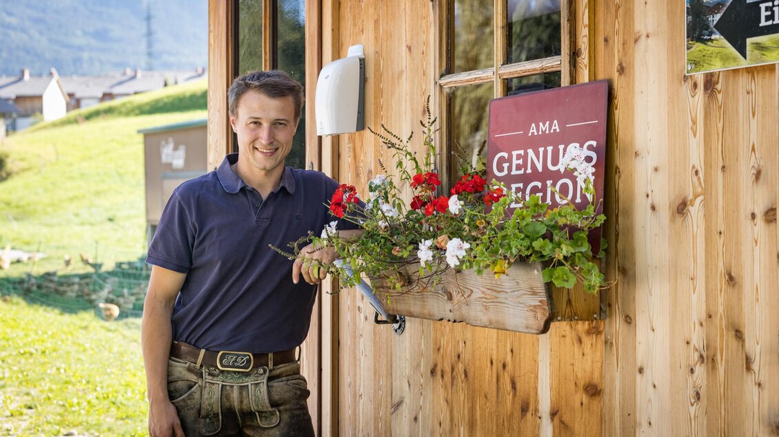 Ein junger Mann steht vor einem Holzgebäude und hält einen Blumenkasten mit bunten Blumen. Im Hintergrund sind grüne Wiesen und Berge sichtbar. | © Netzwerk Kulinarik wildbild