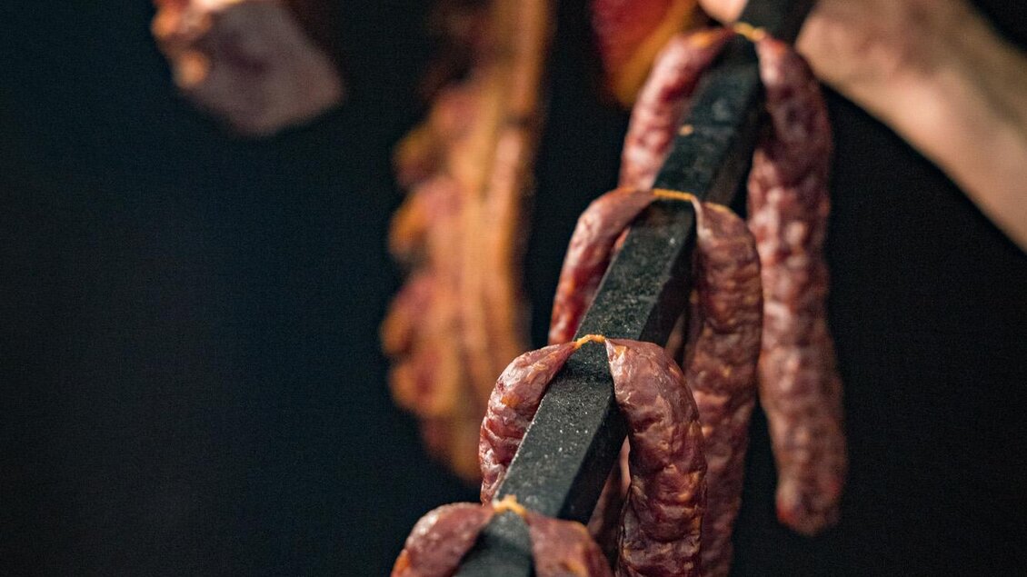 Ein Hand hält eine Stange mit verschiedenen Fleischwaren, darunter Wurst und geräuchertes Fleisch. Im Hintergrund sind weitere Fleischstücke zu sehen. | © Netzwerk Kulinarik wildbild