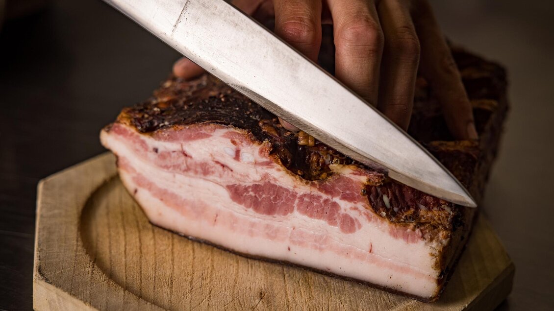 Ein Stück reichhaltiger Speck wird auf einem Holzbrett geschnitten. Ein scharfer Messergriff ist sichtbar, während die Schichtung des Fleisches zur Geltung kommt. | © Netzwerk Kulinarik wildbild