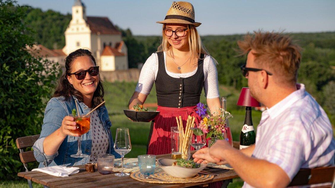 Man sieht zwei draußen sitzende Gäste mit Wein und gutem Essen.  | © Tourismusverband Oststeiermark