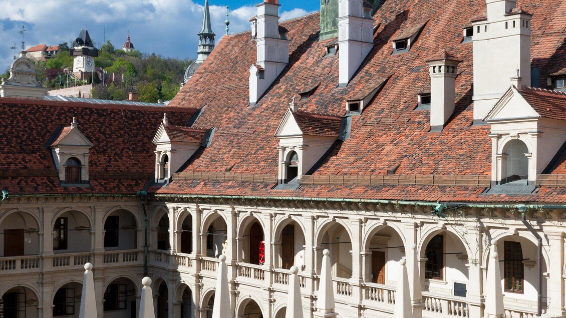 Ein schöner Innenhof eines historischen Gebäudes mit eleganten Arkaden. Die Dächer sind rot gedeckt und die Landschaft im Hintergrund ist grün. | © Graz Tourismus - Harry Schiffer