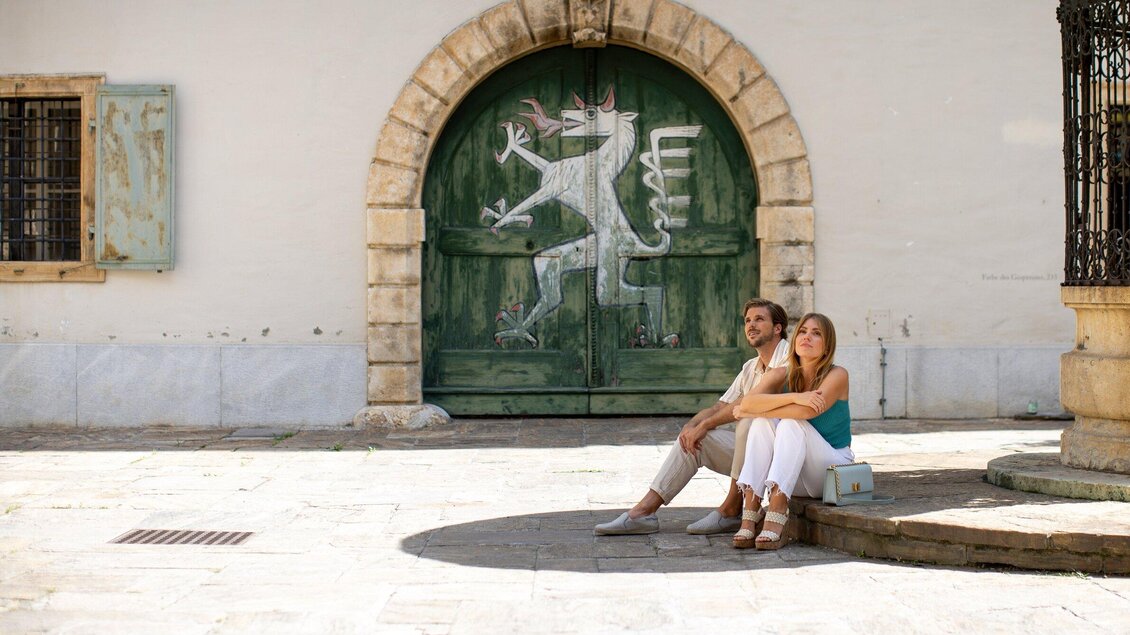 Ein Paar sitzt entspannt vor einer alten grünen Tür mit einem dekorativen Wappen. Die Szenerie ist hell und freundlich, mit einem historischen Gebäude im Hintergrund. | © Graz Tourismus - Tom Lamm