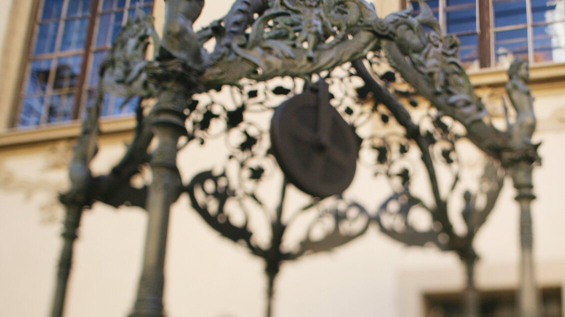Ein kunstvoll gestalteter Brunnen mit einem dekorativen, schmiedeeisernen Gestell. Im Hintergrund sind Fenster eines historischen Gebäudes sichtbar. | © Graz Tourismus - Harry Schiffer