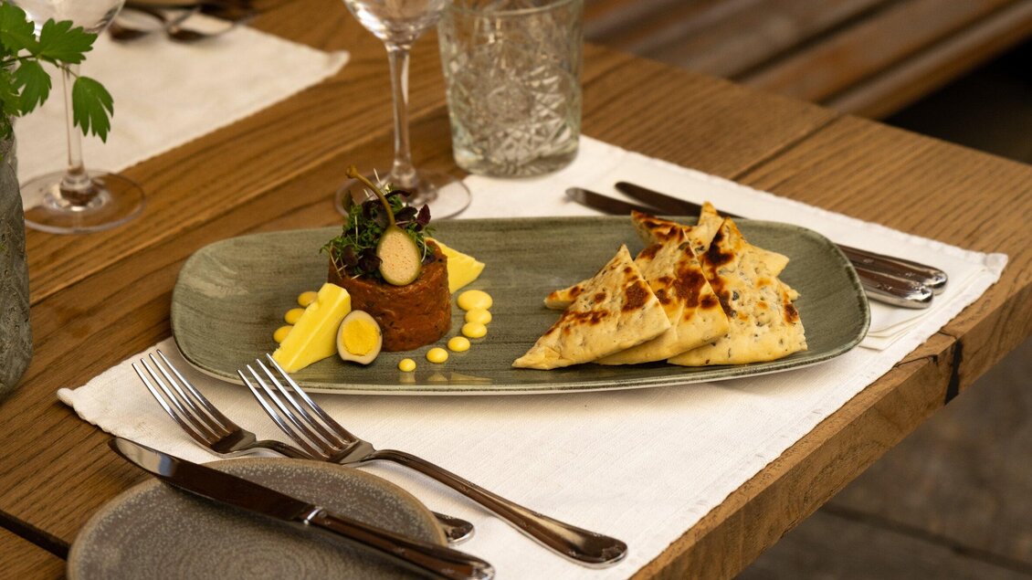 Ein elegantes Gericht auf einem langen Teller, präsentiert mit kunstvollen Elementen. Im Hintergrund sind Gläser und Besteck sichtbar, die eine einladende Tischdekoration bilden. | © Landhauskeller - Benjamin Kern