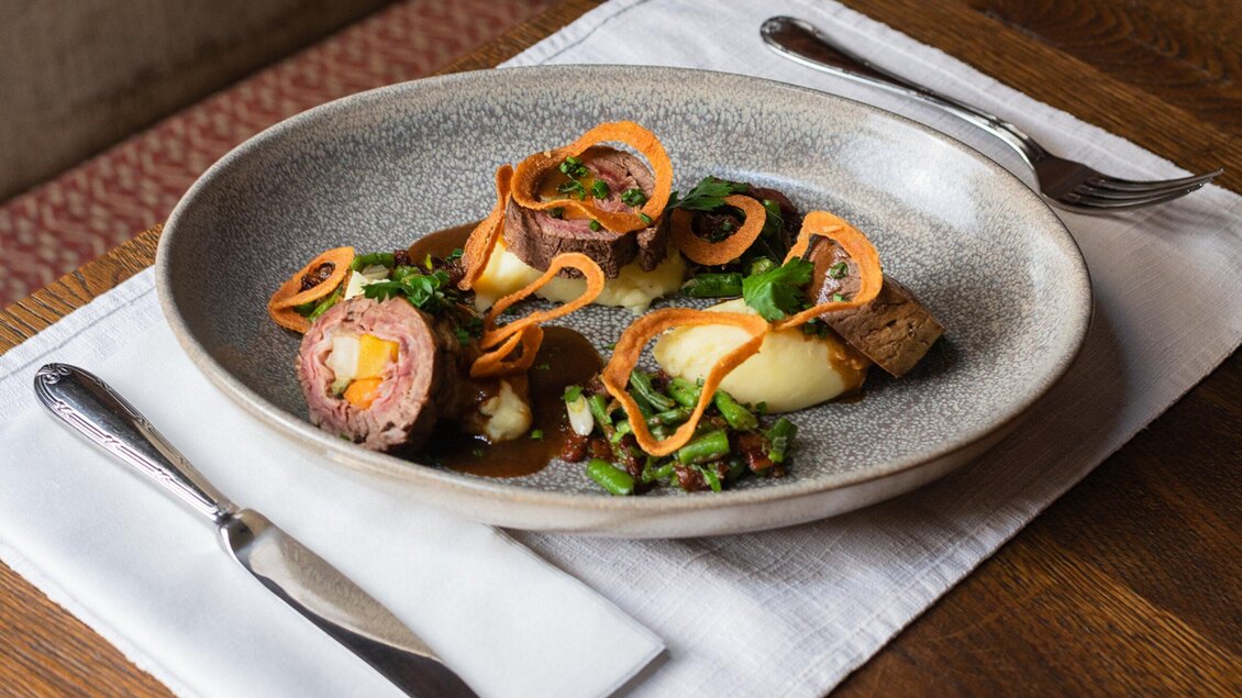 Ein kunstvoll angerichteter Teller mit gefülltem Fleisch, umgeben von pürierten Kartoffeln und frischen Kräutern. Daneben sind knusprige Gemüsestreifen und eine dunkle Sauce sichtbar. | © Landhauskeller - Jauk Katharina