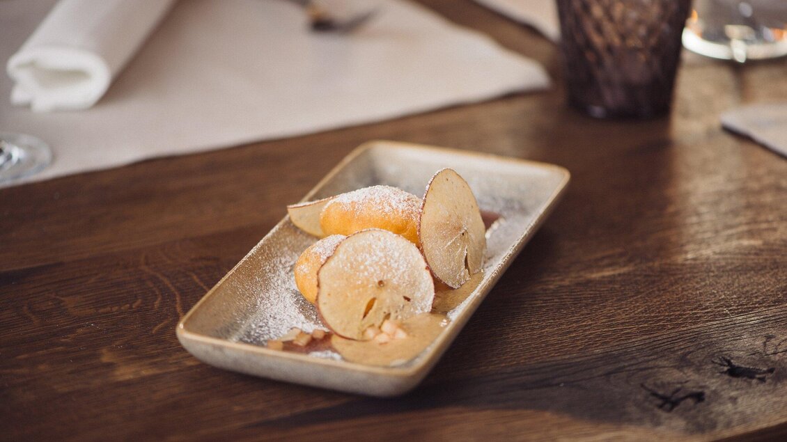 Ein Teller mit süßem Dessert, bestehend aus fruchtigen Scheiben und Puderzucker. Der Hintergrund zeigt eine Holzmaserung und dunkle Dekoration. | © Landhauskeller