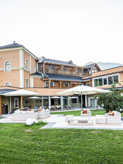 An elegant hotel building surrounded by a green meadow and mountains. The terrace invites you to relax outdoors. | © Landhotel Timmerer