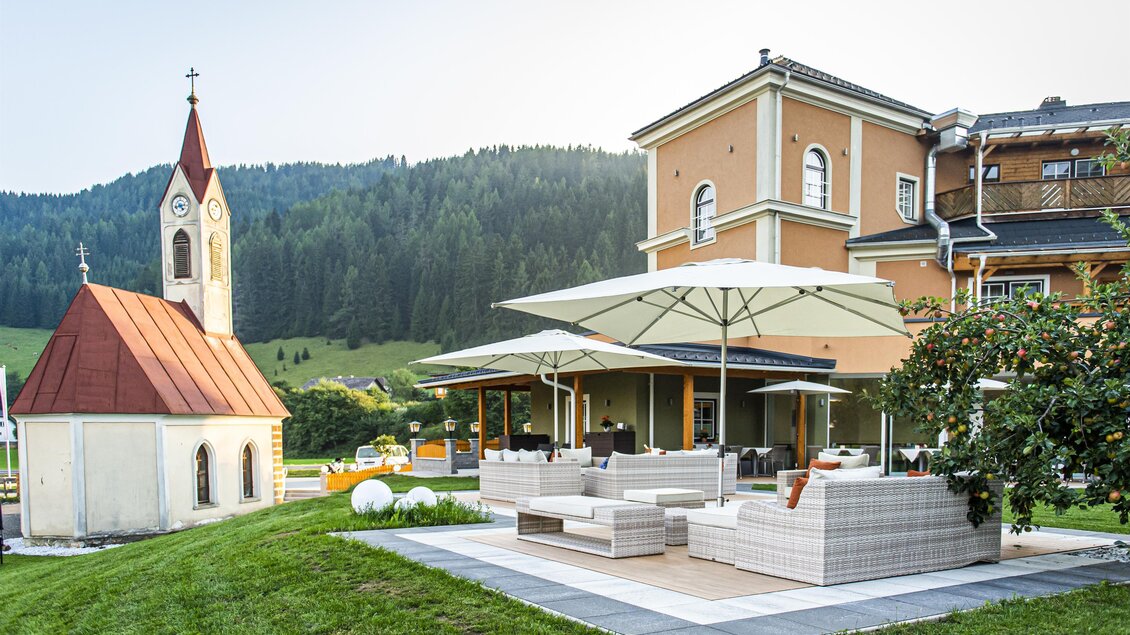 Ein schönes Anwesen mit einer modernen Terrasse und entspannenden Sitzecken. Im Hintergrund steht eine kleine Kirche vor einer malerischen Berglandschaft. | © Landhotel Timmerer