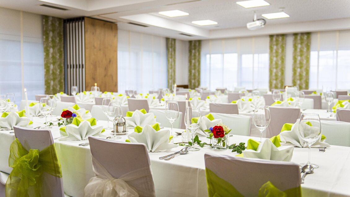 Ein elegant dekorierter Saal mit gedeckten Tischen und gelben Servietten. Die Stühle sind mit grünen Schleifen verziert und große Fenster sorgen für viel Licht. | © Landhotel Timmerer