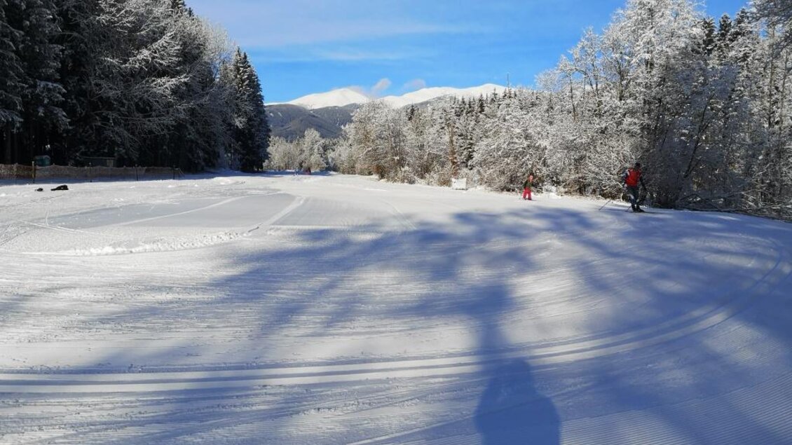 Eine verschneite Landschaft mit schneebedeckten Bäumen und blauem Himmel. Im Hintergrund sind Menschen beim Wintersport zu sehen. | © Anita Fössl