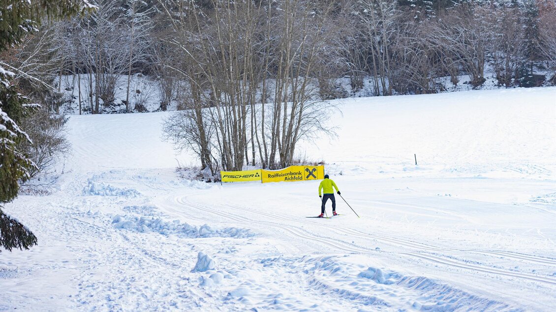 Ein Skilangläufer gleitet durch eine schneebedeckte Landschaft. Im Hintergrund sind schneebedeckte Bäume und eine Werbetafel zu sehen. | © Anita Fössl
