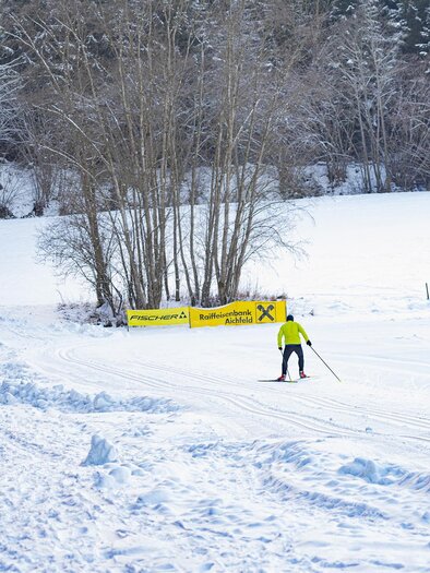 A cross-country skier glides through a snow-covered landscape. In the background, there are snow-covered trees and a billboard visible. | © Anita Fössl