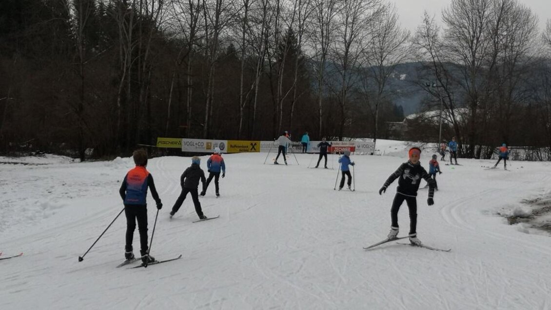 Eine Gruppe von Kindern fährt Skilanglauf auf einer schneebedeckten Fläche. Im Hintergrund sind Bäume und Berge zu sehen. | © Anita Fössl