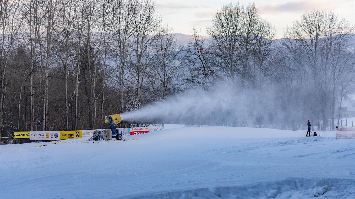 Eine Schneekanone bläst Schnee auf eine glatte, winterliche Piste. Im Hintergrund sind kahle Bäume und ein schneebedeckter Hang zu sehen. | © Anita Fössl