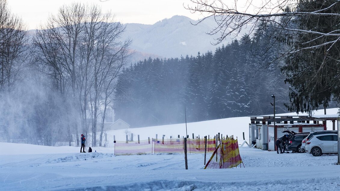 Eine verschneite Landschaft mit Bäumen und Bergen im Hintergrund. Im Vordergrund sieht man ein Skigebiet mit einem Gebäude und einem Parkplatz. | © Anita Fössl