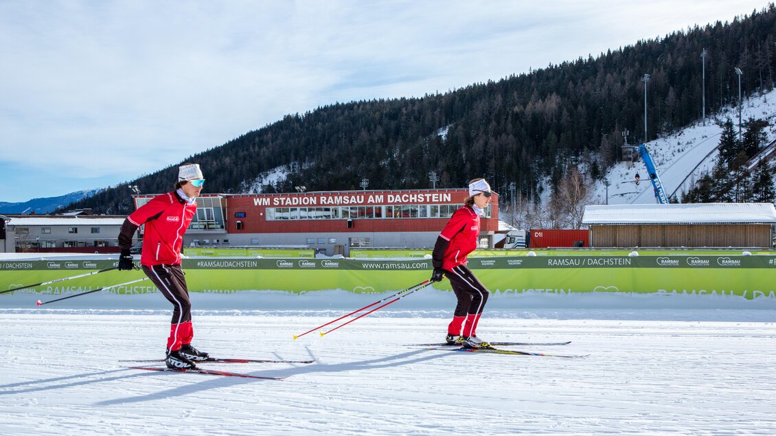 Zwei Skifahrer im rot-weißen Outfit fahren auf einer verschneiten Strecke. Im Hintergrund sind Berge und das Stadion Ramsau am Dachstein zu sehen. | © Alpincenter Dachstein