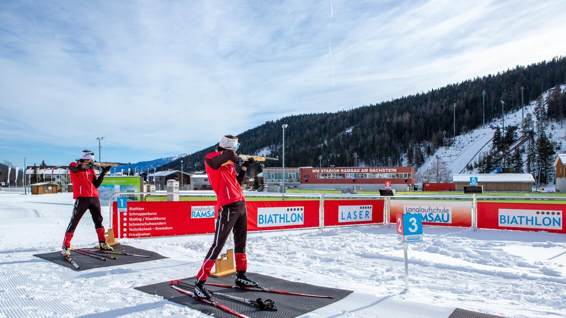 Zwei Skifahrer in roter Kleidung üben in einer schneebedeckten Umgebung. Im Hintergrund sind Berge und ein Biathlon-Areal zu sehen. | © Alpincenter Dachstein