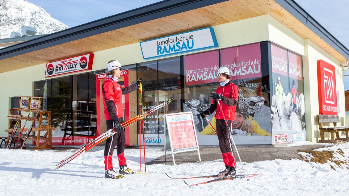 Zwei Skilehrende stehen vor der Langlaufschule Ramsau in einer schneebedeckten Landschaft. Im Hintergrund ist das moderne Gebäude der Schule zu sehen. | © Langlaufbüro | Alpincenter Dachstein
