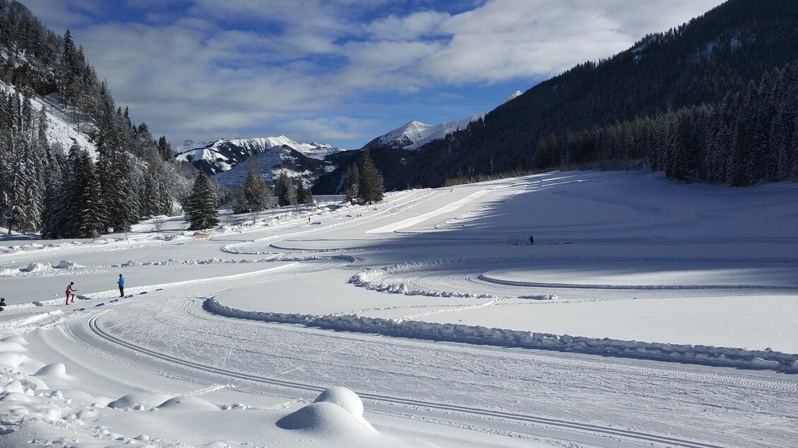 Eine schneebedeckte Landschaft mit Bergen im Hintergrund. Skifahrer sind auf der Strecke unterwegs, während die Sonne am Himmel scheint. | © NAZ Erzberg Arena