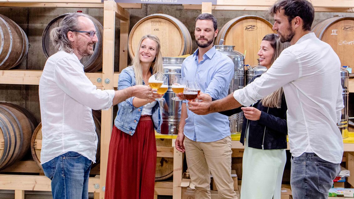 Eine Gruppe von vier Personen hebt Gläser mit Getränken in einer Brauerei an. Im Hintergrund sind Holzfässer und metallische Behälter zu sehen. | © Lava Bräu