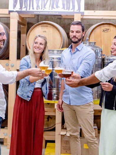 A group of four people is raising glasses with drinks in a brewery. In the background, wooden barrels and metallic containers can be seen. | © Lava Bräu